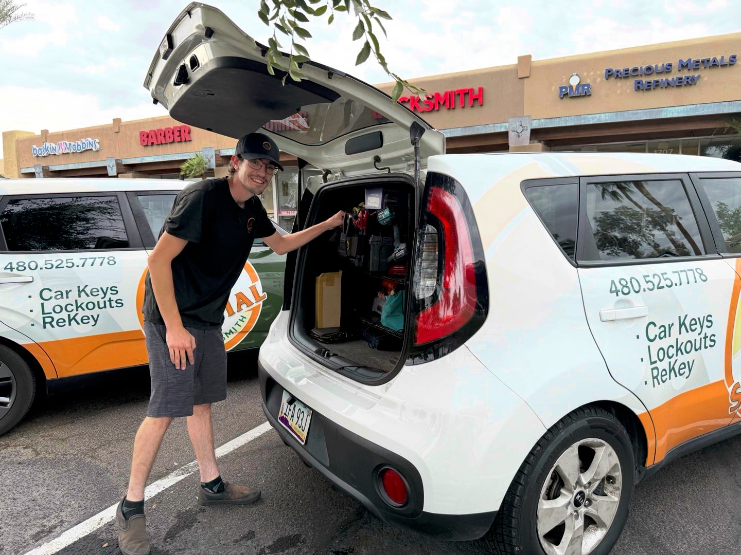 A locksmith technician stands beside an open hatchback car branded with Car Keys Lockouts ReKey signage in a parking lot.