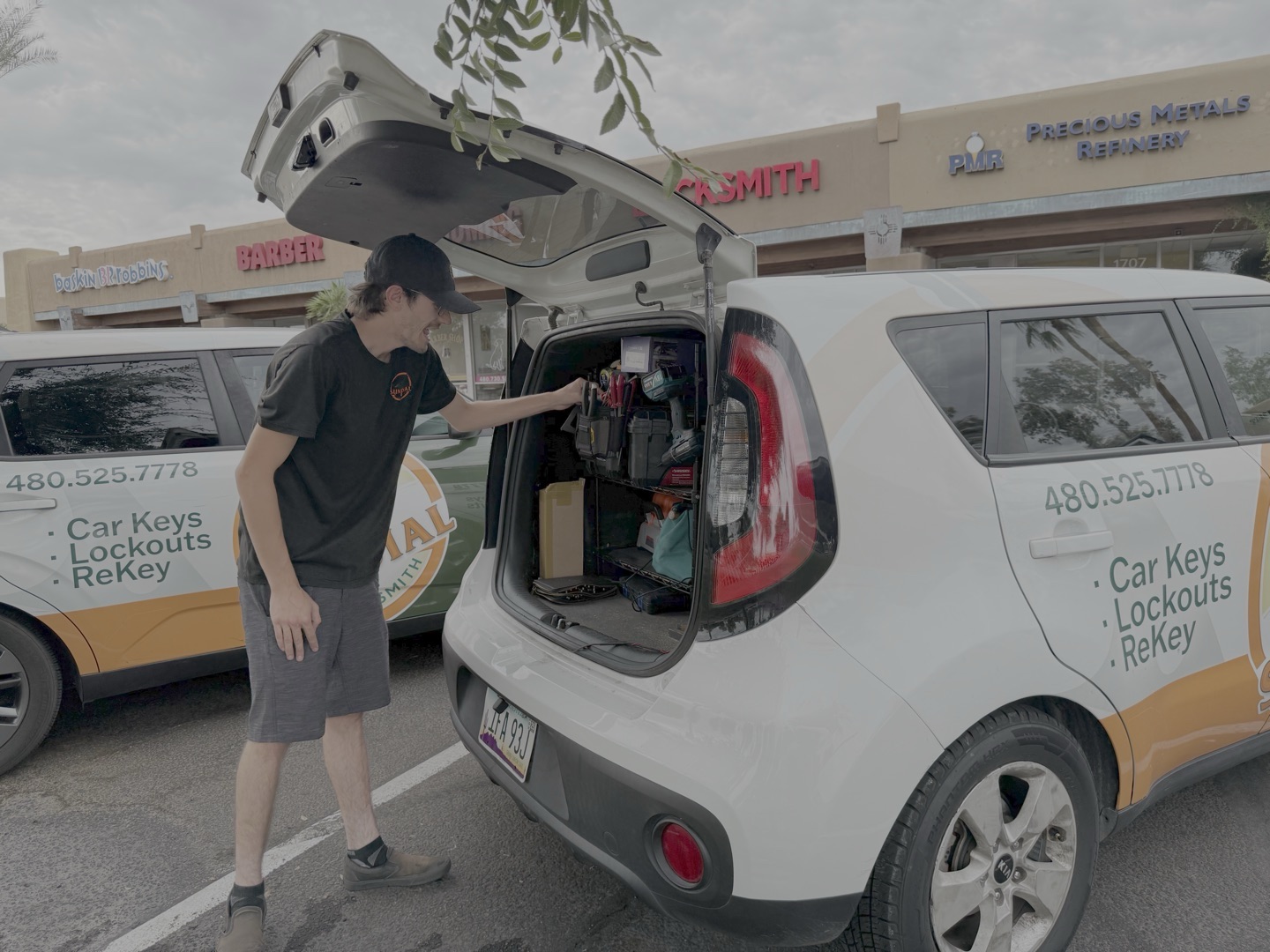 A locksmith technician in a black uniform opens the rear hatch of a branded white Kia Soul service vehicle stocked with tools and supplies in a strip mall parking lot.