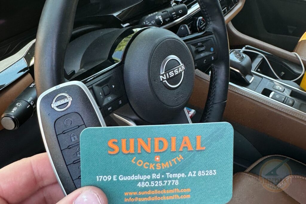 A hand holding a Nissan smart key fob and a Sundial Locksmith business card inside a Nissan vehicle with the steering wheel visible in the background.
